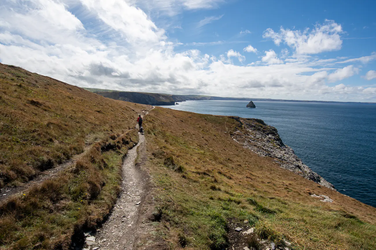 A walker on a narrow coastal path above the sea, with headlands stretching into the distance