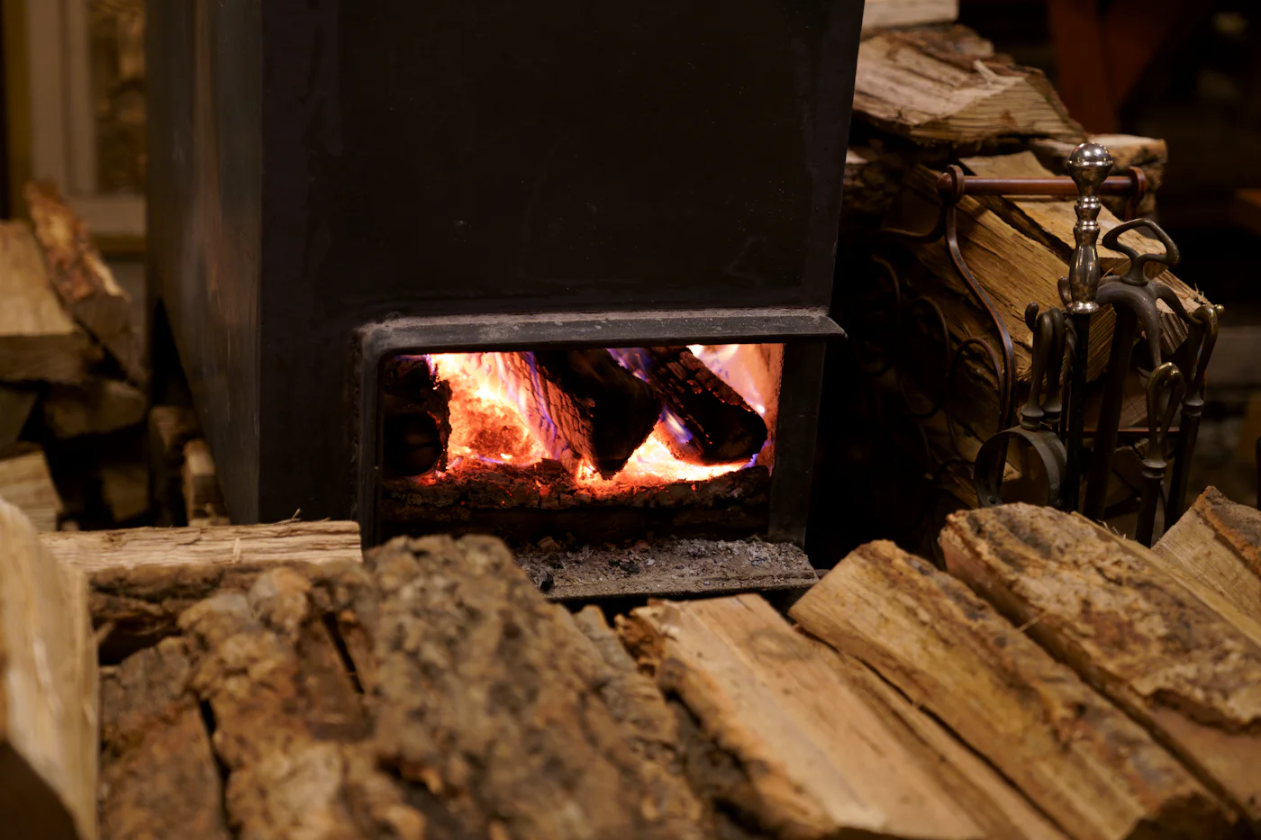 A black wood-burning stove glowing beside a stack of logs